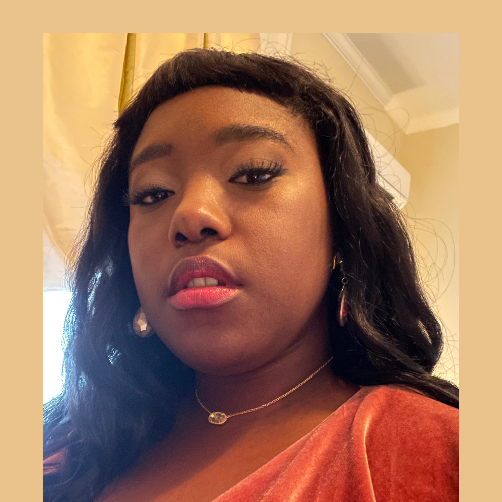 Portrait of a woman with medium brown skin and long dark hair wearing an orange top, photographed indoors with soft lighting and a neutral background.