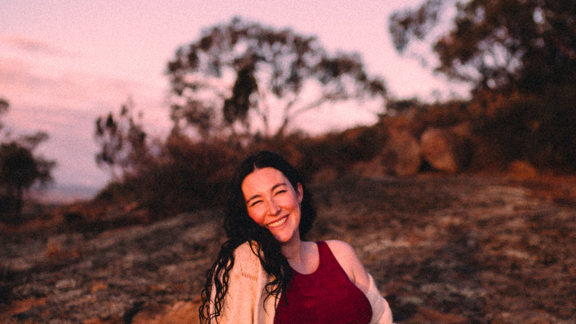 A woman, Isabella Frappier, with long dark hair smiles warmly while standing outdoors in soft evening light, wearing a sleeveless deep red dress with trees and sky blurred in the background.