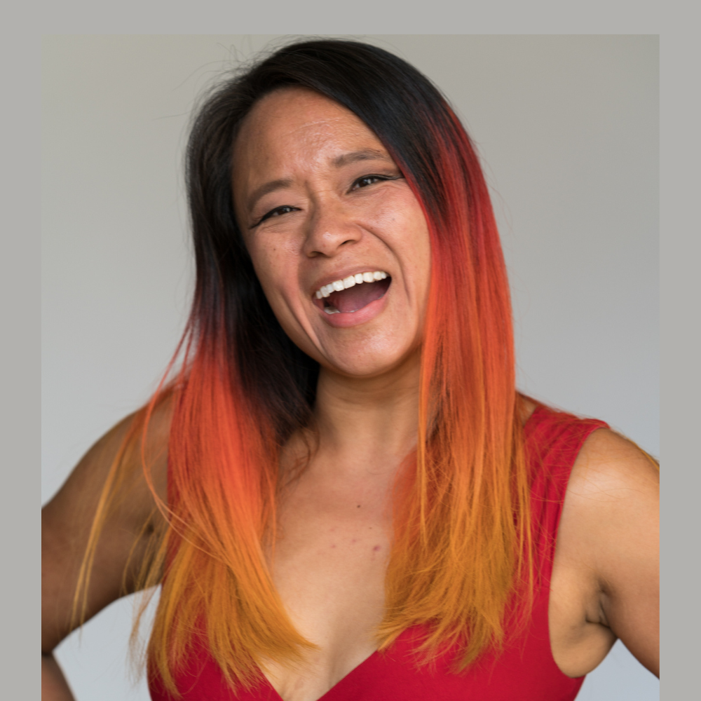 Portrait of a woman with long dark hair and red highlights wearing a sleeveless top, smiling broadly against a plain studio background.