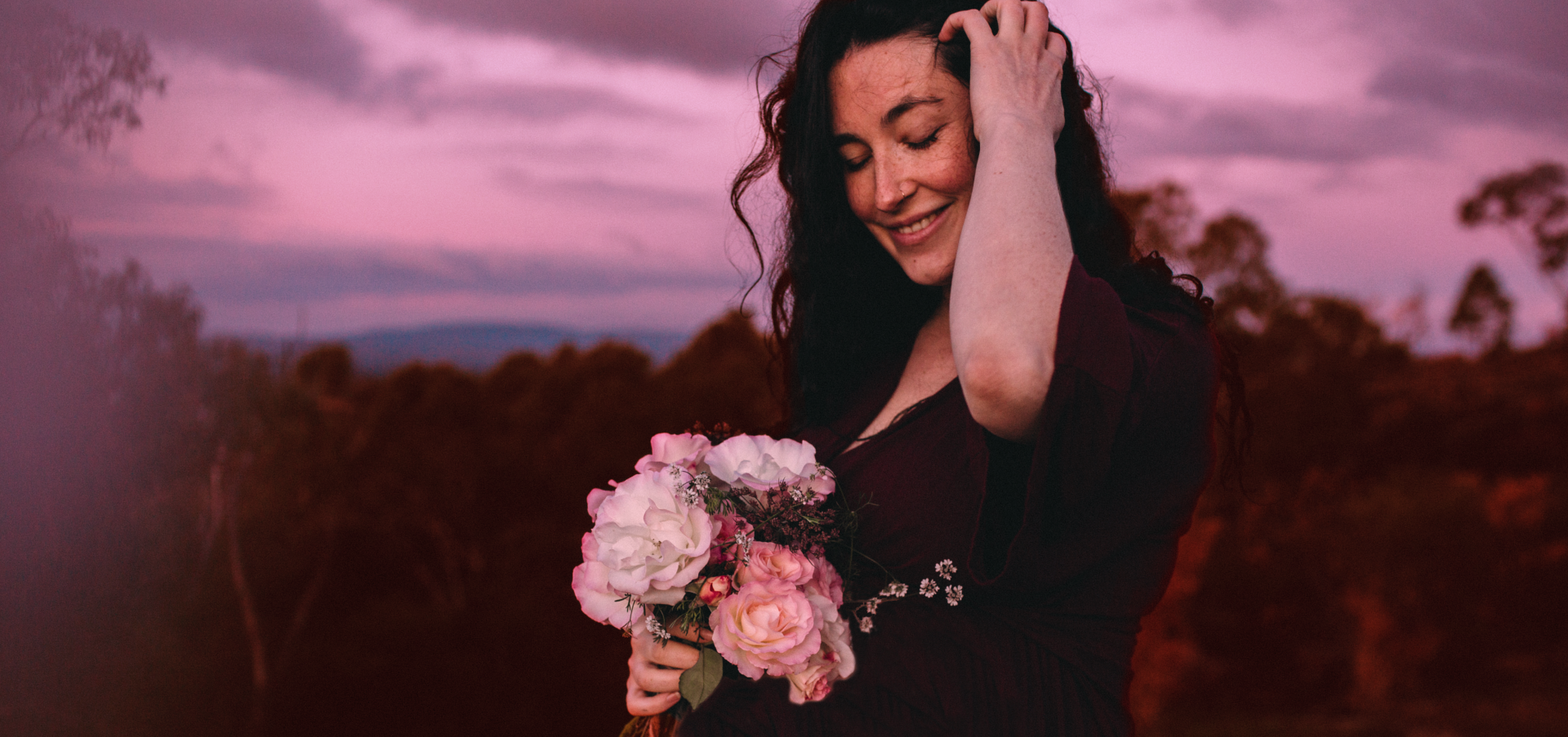 Isabella Frappier, pleasure mentor and founder of The Pleasure Sanctuary, a woman with long dark hair wearing a deep red dress holds a bouquet of pink roses outdoors at dusk, smiling gently in warm natural light.